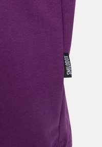 Primo piano di un tessuto di cotone viola, caratterizzato da una texture a coste e un'etichetta nera con testo bianco sul lato.