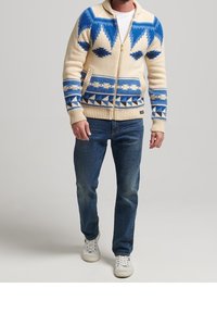 Pletená svetr zip-up v krémové barvě s modrými geometrickými vzory, disponující límečkem, bočními kapsami a žebrovanými manžetami. V kombinaci s modrými džíny.