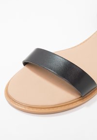 Mustat nahkasandaalit, joissa on leveä hihna beige pohjan päällä. Sandalit ovat sileäpintaiset ja niissä on litteä pohja, jonka reunassa on ompeleet.