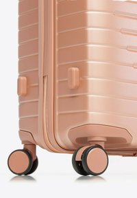 Valise rigide en or rose avec une texture rainurée, deux roulettes à mouvement fluide et fermeture éclair. Comprend des poignées latérales robustes.