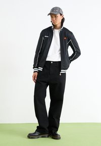 Sort nylonjakke med hvide striber, sorte brede bukser og sorte snørebods støvler. Grå kasket med cirkulært mønster. Står foran en hvid baggrund.