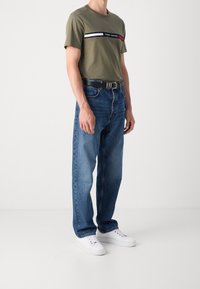 Muž v olivově zeleném tričku Tommy Hilfiger, s modrými džíny a černým opaskem, a bílými teniskami, stojí před jednobarevným světlým pozadím.