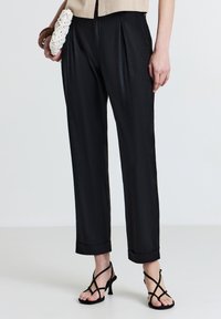 Pantaloni neri plissettati con una leggera lucentezza, vestibilità sartoriale, orlo a risvolto, abbinati a tacchi neri con cinturini e una pochette bianca strutturata.