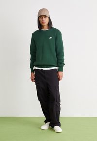 Πράσινη πλεκτή μπλούζα με λευκό λογότυπο Nike, συνδυασμένη με μαύρο cargo παν τελόνι. Λευκά αθλητικά παπούτσια και μπεζ καπέλο ολοκληρώνουν το σύνολο.