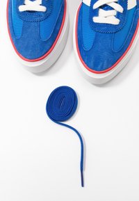 Blue suede and fabric sneakers with white laces, featuring a red sole trim. A blue shoelace is coiled beside the shoes on a white background.