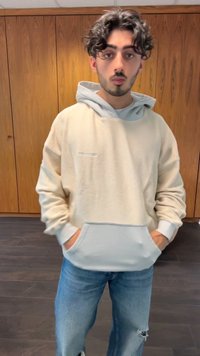 Svetlo bež oversized kapučo s svetlejšo sivo kapuco in sprednjim žepom, izdelano iz teksturirane tkanine. Nosene so z raztrganimi modrimi džins hlačami.