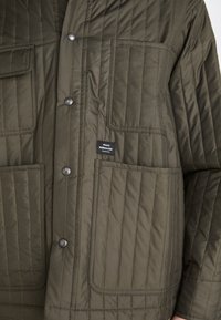 Quilted Jacke in olivgrün mit Druckknöpfen, zwei Fronttaschen und einem Etikett. Glatte Textur mit vertikalen Nahtdetails.