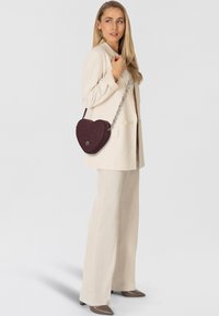 Bolso burdeos en forma de corazón con patrón en relieve, con una correa de cadena plateada y textura de cuero suave. Llevado sobre un blazer color crema.