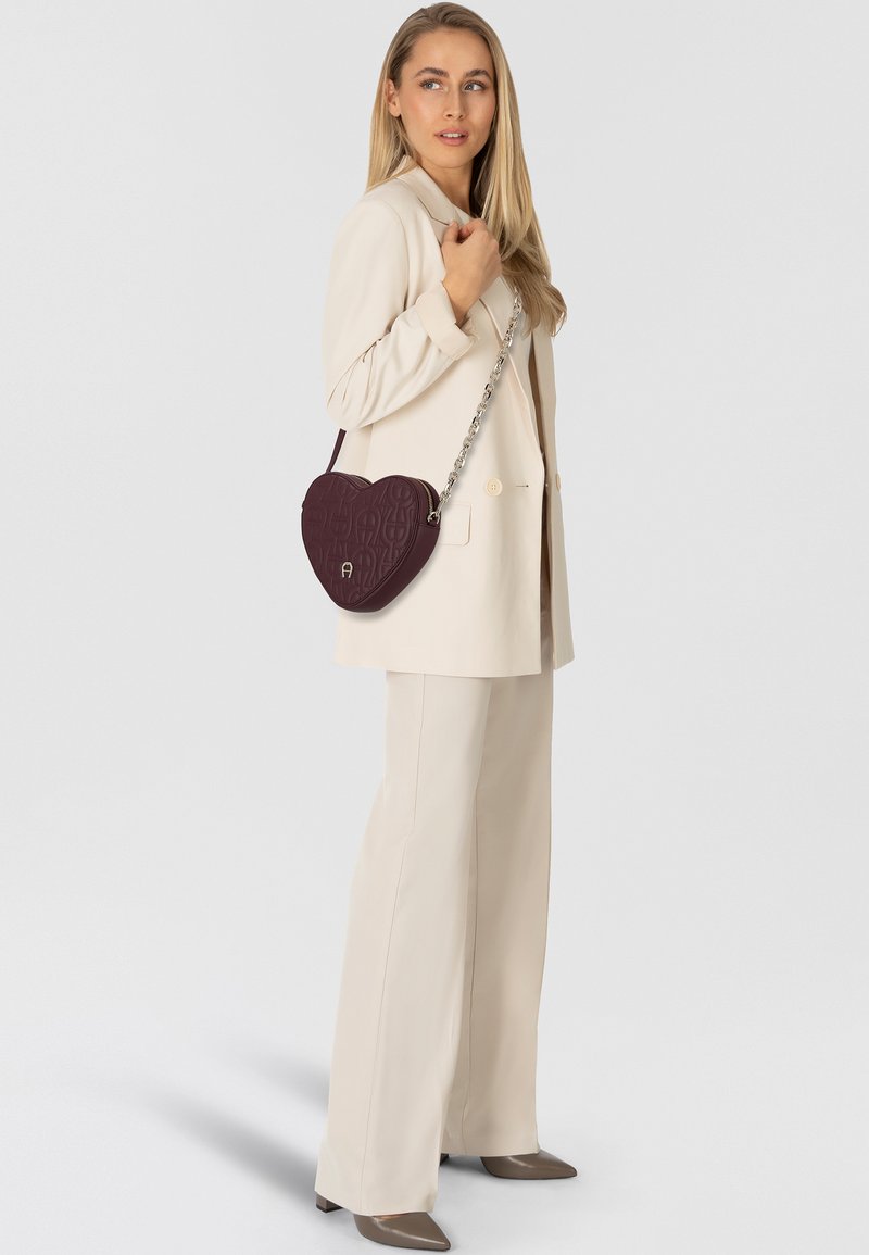 Bolso burdeos en forma de corazón con patrón en relieve, con una correa de cadena plateada y textura de cuero suave. Llevado sobre un blazer color crema.