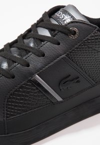 Svart lädersneaker med strukturerade detaljer, tonade snören och ett silverstreck. Har en krokodillogo och en slät, blank yta.