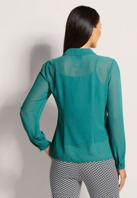 Türkisfarbene, durchsichtige Bluse mit langen Ärmeln und Kragen, die an den Schultern gerafft ist und unten einen strukturierten Fischgrätenmuster aufweist.