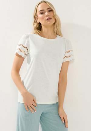 Vaaleahiuksinen nainen, jolla on valkoinen lyhythihainen yläosa, jossa on leikattuja kuvioita, ja vaaleansiniset housut, seisomassa yksinkertaisen valkoisen taustan edessä.