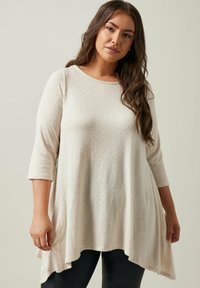 Tunica beige e leggera con maniche a 3/4, texture sottile e motivo a pois. L'orlo è asimmetrico, creando una silhouette fluida.