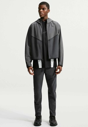 Giovane uomo in piedi, indossa una giacca con cappuccio e zip grigio scuro, maglietta sportiva nera con accenti bianchi, pantaloni abbinati e sneakers nere.
