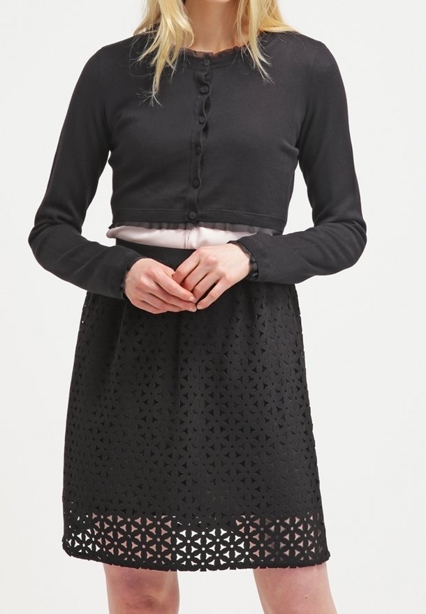 Cardigan noir court avec boutons, associé à une robe noire présentant des motifs triangulaires découpés. Les deux ont des manches longues et une silhouette ajustée.
