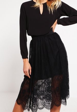 Blusa nera a maniche lunghe con scollo a chiave abbinata a una gonna midi in pizzo nero. La gonna presenta un bordo scallopato e una texture in pizzo a strati.