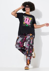 T-shirt nera con una grande grafica di un maiale rosa, abbinata a pantaloni colorati a fantasia con vari disegni e testi. Sandali con una fascia bianca.