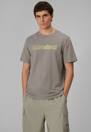 Jonge man met kort donker haar, draagt een grijs T-shirt met gele tekst en lichtbeige cargoshorts, staat met zijn handen in zijn zakken.