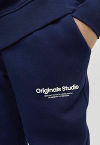 Tumši zila kokvilnas sporta jaku ar priekšējo kabatu, ribotām piedurknēm un baltu logo tekstu "Originals Studio" apakšējā labajā stūrī.