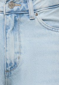 Détail d'un jean en denim bleu clair montrant la poche avant, la passants de ceinture et le bouton métal avec des accents de couture.