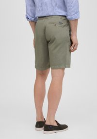 Los pantalones cortos caqui presentan un corte estándar, un bolsillo trasero con cierre de botón y una cinturilla cómoda. Se llevan con una camisa a rayas azul y unos zapatos oscuros sin cordones.