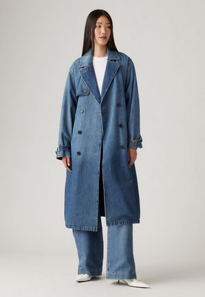 Lång denimtrenchcoat med dubbelknäppt design, ljus till medeltvätt, stora slag och svarta knappar. Matchas med utsvängda jeans.