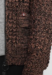 Cardigan lavorato a maglia marrone e nero con pattern testurizzato, chiusura con zip e una piccola patch logo sulla tasca. Costruzione in maglia spessa e accogliente.