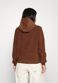 Sudadera con capucha de vellón marrón con acabado texturizado, que cuenta con una capucha a juego y un dobladillo acanalado, usada sobre pantalones de color crema.