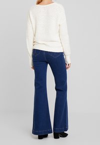 Jasny kremowy sweter z dzianiny o luźnym, teksturowanym wzorze, w połączeniu z ciemnoniebieskimi spodniami dżinsowymi w kształcie dzwonów z detalicznymi tylnymi kieszeniami.