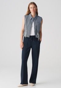 Gilet in denim senza maniche con righe blu e bianche, vita aderente con cordini, abbinato a pantaloni blu navy e scarpe beige.