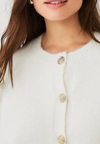 Witte gebreide cardigan met een ronde halslijn, voorzien van vier gestructureerde gouden knopen en een zachte, licht pluizige textuur.