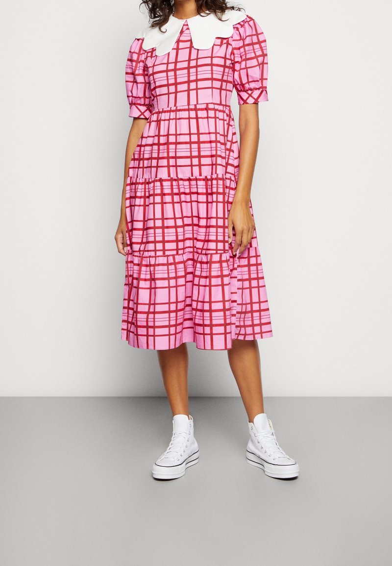 Femme portant une robe midi à carreaux rose et rouge avec des manches bouffantes, un grand col blanc et des baskets montantes blanches sur un sol gris.