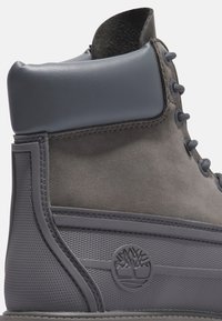 Bottine en suède gris avec un embout rond, cuir à l'extérieur, lacets gris et panneau texturé. Comprend un logo en relief sur le côté.