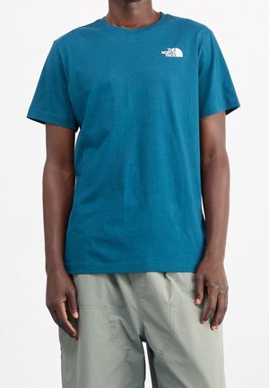 Mand iført en kortærmet The North Face t-shirt i teal og lysegrå bukser, stående mod en ensfarvet hvid baggrund.