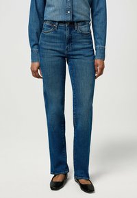 Mujer con jeans rectos de tiro medio en azul, una camisa de mezclilla a juego y zapatos planos negros, de pie contra un fondo liso.