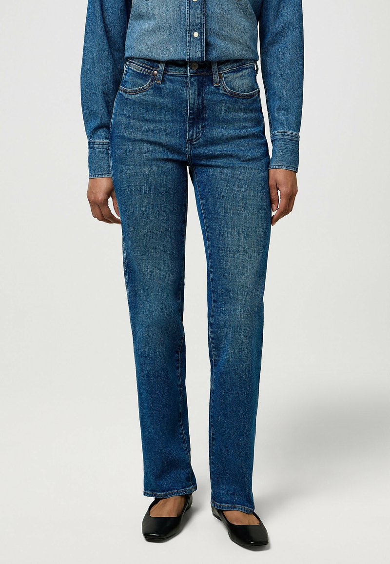 Mujer con jeans rectos de tiro medio en azul, una camisa de mezclilla a juego y zapatos planos negros, de pie contra un fondo liso.