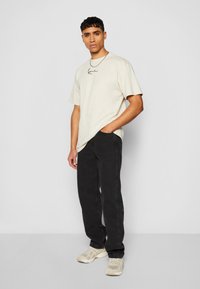 Beige cotton t-shirt with black graphic text, paired with black jeans. The model wears light-colored sneakers, standing against a gray background.