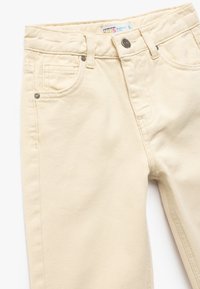 Jeans in cotone beige per bambini con vestibilità comoda, dotati di cinque tasche, chiusura con bottone e cerniera, e dettagli di cucitura sottili.