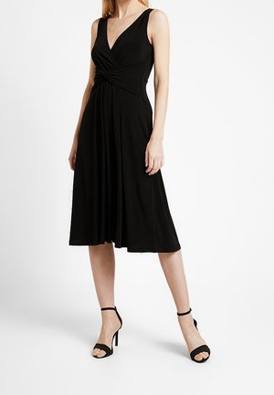 Kobieta ubrana w czarną midi sukienkę bez rękawów z marszczonym węzłem w talii, w połączeniu z czarnymi szpilkami na kostce.