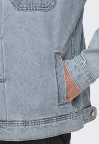 Ljust blå denimjacka med en ficka, synliga sömmar och en något blekt textur. Ärmarna är uppvikta.