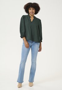 Blusa verde scuro texture con scollo a V e maniche a palloncino, abbinata a jeans a zampa d'elefante blu chiaro e stivali con tacco beige.