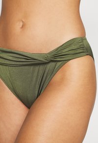 Olivengrønn bikinitruse laget av glinsende materiale, med et tvunnet frontdesign og innsamlet detaljer langs sidene.