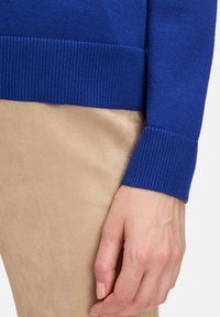 Blå stickad tröja med ribbade manschetter, i kombination med beiga mockalika byxor. Fokusera på texturen och färgkontrasten mellan plaggen.