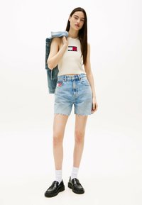 Lyst bundet mørkeblå denimshorts med en frynset kant, parret med en ribbet hvid crop top med et rødt og blåt logo. Sorte loafers og en denimjakke.