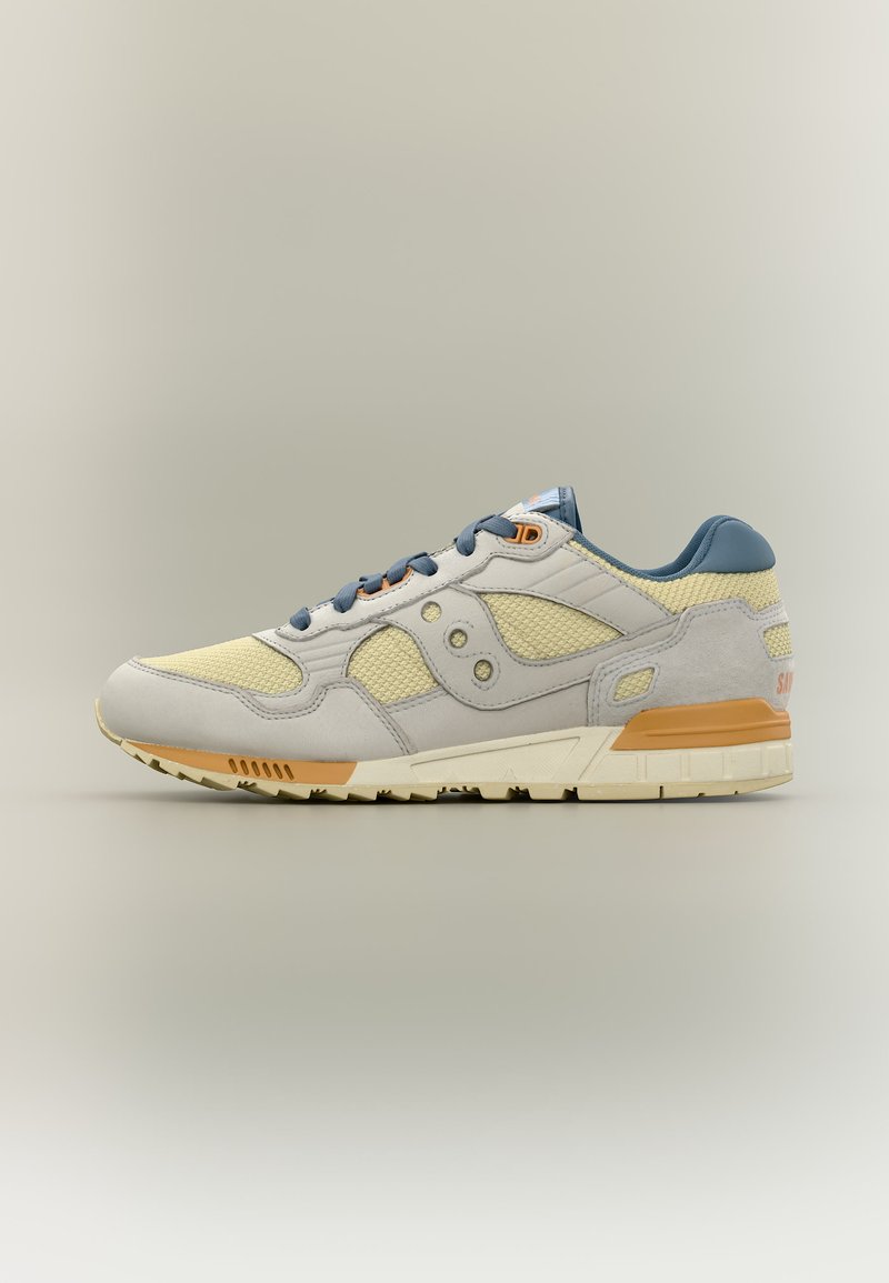 Baskets beige et gris clair avec panneaux en mesh et daim, lacets bleus et accents orange sur une semelle blanche épaisse, vues de profil.