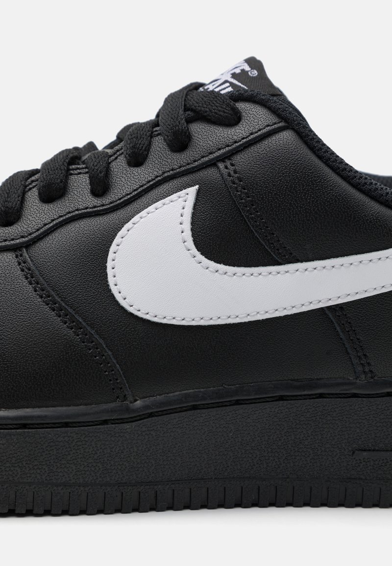 Sneaker en cuir noir avec un logo Nike blanc. Présente une texture lisse, des lacets plats et une semelle en caoutchouc avec des rainures horizontales.