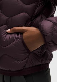 Burgundowa kurtka puchowa z teksturowanym pikowanym wzorem. Zawiera dłoń w kieszeni. Gładki, błyszczący materiał z kontrastową taśmą u dołu.