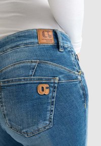 Jeans en denim bleu moyen. Comprend une patch en cuir avec un logo, des coutures contrastantes et deux poches arrière avec des coutures courbées.