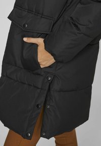 Melna pūkainā jaka ar pogu aizdari un izmatotu dizainu. Gluda auduma un nedaudz spīdīga tekstūra. Aizvērta apakšdaļa.