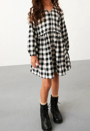 Robe à carreaux noir et blanc avec un col en V, des manches longues et des boutons en bois, associée à des bottes noires à lacets et des chaussettes côtelées beiges.